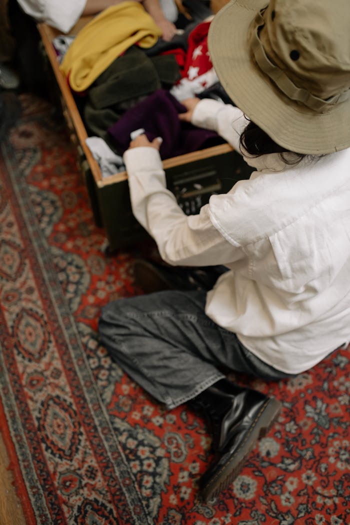 Person in hat sorting clothes in a vintage wooden box on a patterned rug indoors.